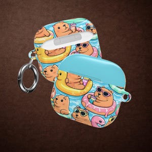 Puede incluir: Dos fundas protectoras para auriculares inalámbricos con un patrón repetitivo de carpinchos de dibujos animados flotando en flotadores de colores sobre agua azul. Los carpinchos llevan gafas de sol y sostienen bebidas. Una funda incluye una anilla.