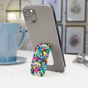Könnte beinhalten: Ein Smartphone mit einem farbenfrohen Pop-up-Telefonhalter im Retro-80er-Design. Der Halter hat einen weißen Ständer und ist mit geometrischen Formen in Pink, Gelb, Blau und Schwarz verziert. Das Telefon ist grau und liegt auf einer weißen Oberfläche.
