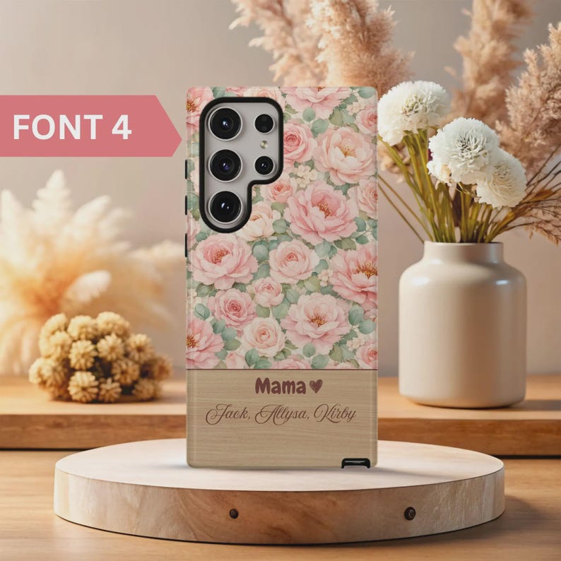 Pode incluir: Capa de telefone floral com rosas cor-de-rosa e folhas verdes. A parte inferior tem um aspeto de madeira com a palavra "Mama" e tr&ecirc;s nomes. O texto "FONT 4" &eacute; vis&iacute;vel no canto superior esquerdo.