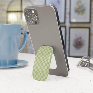 Könnte beinhalten: Ein silbernes Smartphone mit einem grün-weiß karierten Handyhalter steht auf einer weißen Oberfläche. Eine blaue Tasse und Untertasse befinden sich im Hintergrund. Gerahmte Kunst hängt an der Wand.