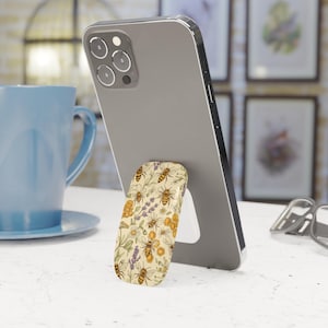 May include: A smartphone is propped up by a beige phone grip with a bee and floral pattern. The grip features illustrations of bees, lavender, and honeycomb. The phone is gray, and a blue mug is in the background.