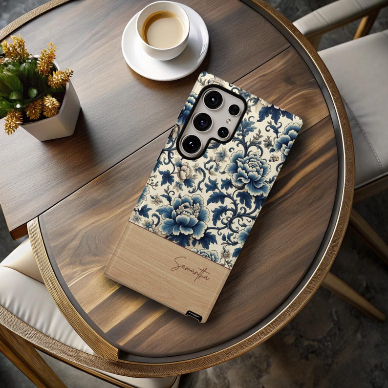 Pode incluir: Uma capa de smartphone com um design floral em azul e creme, e uma se&ccedil;&atilde;o inferior de madeira. O nome "Samantha" est&aacute; escrito em it&aacute;lico na se&ccedil;&atilde;o de madeira. A capa est&aacute; em uma mesa de madeira com uma x&iacute;cara de caf&eacute;.