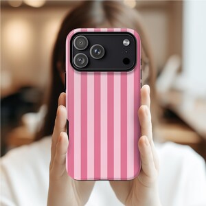 May include: A pink and light pink striped phone case with a black camera module. The case is held up in front of a person. The case has a vertical stripe pattern.