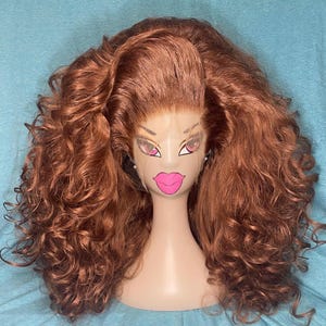May include: A brown wig with voluminous, curly hair styled on a mannequin head. The mannequin has a face with pink lips and eye makeup. The wig is a reddish-brown color and has a lace front.