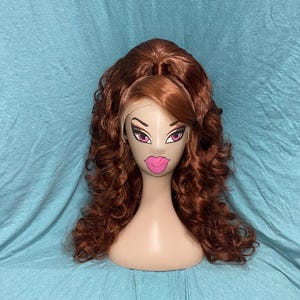 Könnte beinhalten: Ein rotbrauner Perücke mit langen, lockigen Haaren wird auf einem Schaufensterkopf präsentiert. Die Schaufensterpuppe hat ein cartoonartiges Gesicht mit großen Augen und rosa Lippen. Die Perücke hat eine Spitze vorne und einen Seitenscheitel.