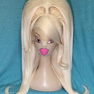 Può includere: Parrucca bionda lunga con coda di cavallo alta e punte arricciate. La parrucca è esposta su una testa di manichino con labbra rosa e trucco occhi, su sfondo blu.