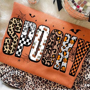 May include: Orange Halloween-themed sweatshirt with the word "SPOOKY" in a variety of patterns, including leopard print, checkered, and pumpkin designs. The design includes bats, spiderwebs, and stars.