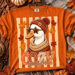 May include: An orange t-shirt featuring a graphic of a white duck wearing a rust-coloured knit beanie with a small pumpkin icon, a matching knit scarf, and star-shaped sunglasses. The text COZY season is printed below the duck illustration.