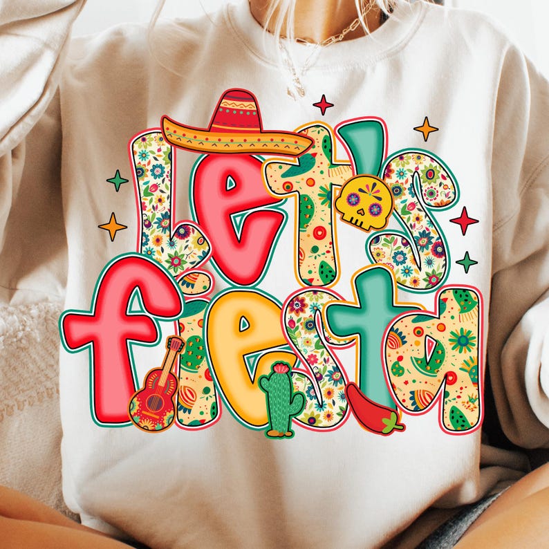 May include: A cream-colored sweatshirt with a colorful "Let's Fiesta" graphic. The design includes a sombrero, guitar, cactus, chili pepper, and a skull, all in vibrant colors and floral patterns. The text is outlined in black.