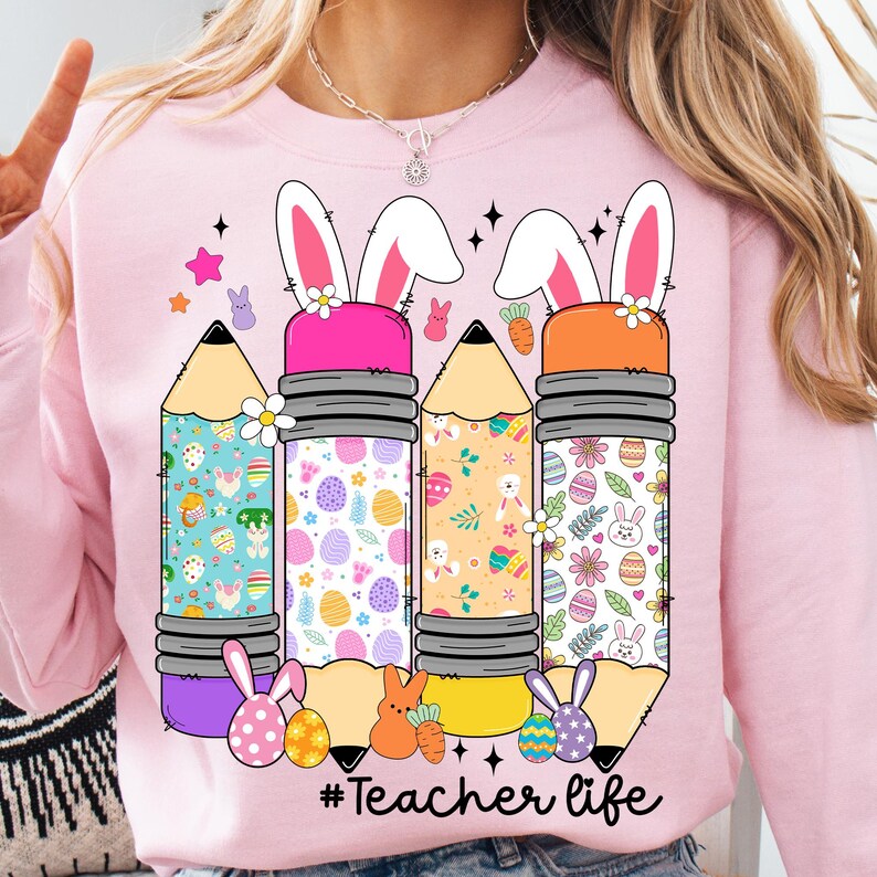 Op de afbeelding: Roze trui met een afbeelding van vier potloden met konijnenoren en paasei-patronen. De tekst "#Teacher life" is onder de potloden gedrukt.