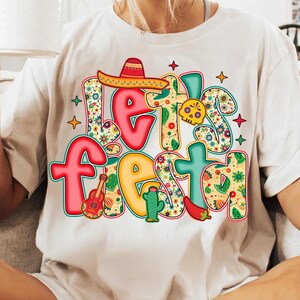 May include: White t-shirt with the words "Let's Fiesta" in colorful, festive lettering. The design includes a sombrero, guitar, cactus, chili pepper, and a skull, all in vibrant colors and floral patterns, perfect for a celebration.
