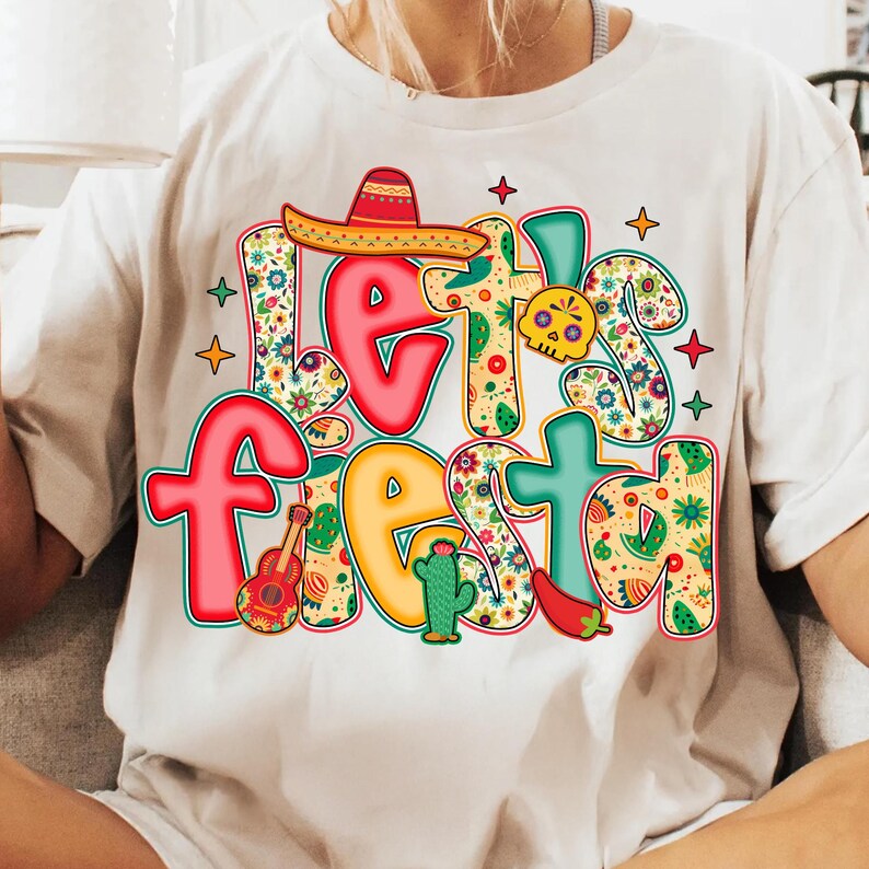 May include: White t-shirt with the words "Let's Fiesta" in colorful, festive lettering. The design includes a sombrero, guitar, cactus, chili pepper, and a skull, all in vibrant colors and floral patterns, perfect for a celebration.