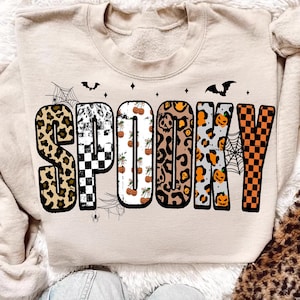 May include: Beige sweatshirt with the word "SPOOKY" in a variety of patterns, including leopard print, checkerboard, and pumpkins. The design includes bats, spiderwebs, and stars, perfect for Halloween.