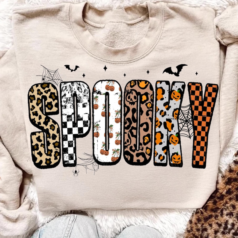 May include: Beige sweatshirt with the word "SPOOKY" in a variety of patterns, including leopard print, checkerboard, and pumpkins. The design includes bats, spiderwebs, and stars, perfect for Halloween.