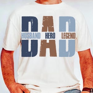 以下が含まれることがあります： クリーム色のTシャツ。青と茶色のブロック体で「DAD」と「DAD」の文字がプリントされています。「HUSBAND」、「HERO」、「LEGEND」の文字も同様のスタイルでプリントされています。カジュアルな普段使いのTシャツ。