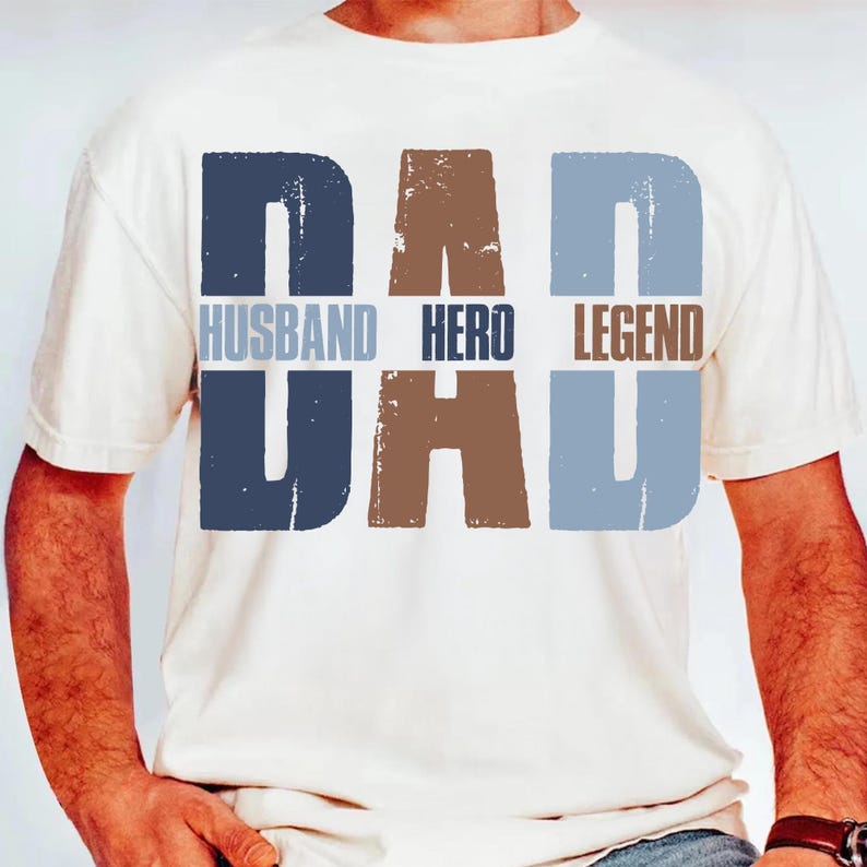 以下が含まれることがあります： クリーム色のTシャツ。青と茶色のブロック体で「DAD」と「DAD」の文字がプリントされています。「HUSBAND」、「HERO」、「LEGEND」の文字も同様のスタイルでプリントされています。カジュアルな普段使いのTシャツ。