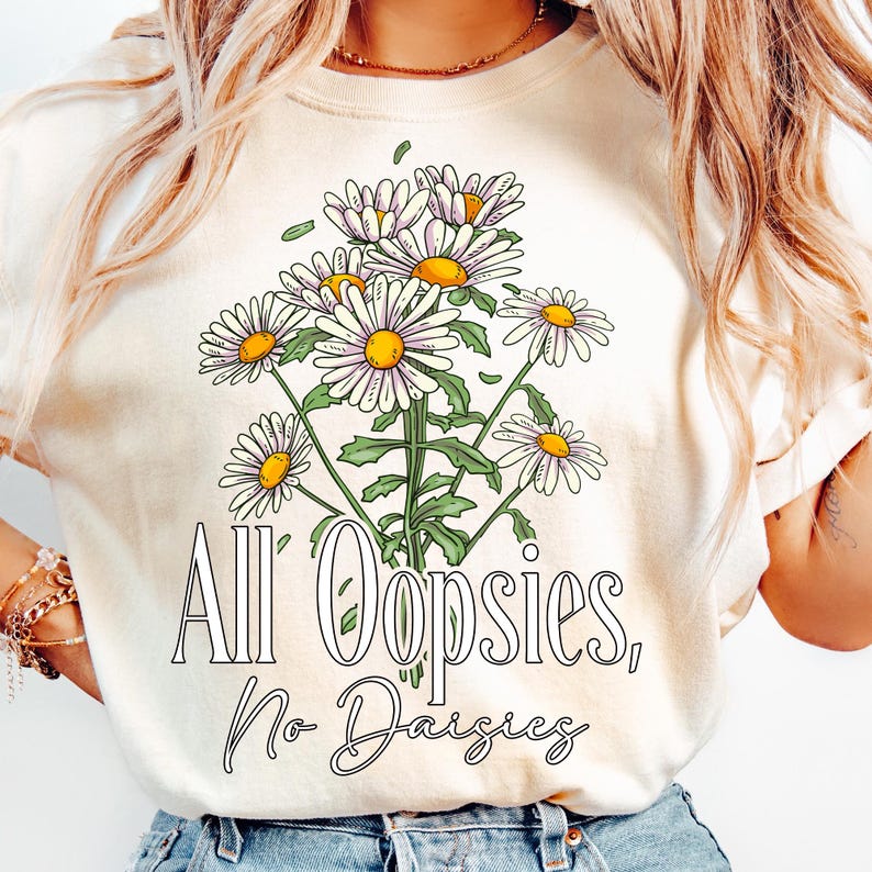 K&ouml;nnte beinhalten: Cremefarbenes T-Shirt mit einem Bouquet aus G&auml;nsebl&uuml;mchen mit wei&szlig;en Bl&uuml;tenbl&auml;ttern und gelben Zentren, gr&uuml;nen Stielen und Bl&auml;ttern. Der Text "All Oopsies, No Daisies" ist unter dem floralen Design in einer Schriftart gedruckt.