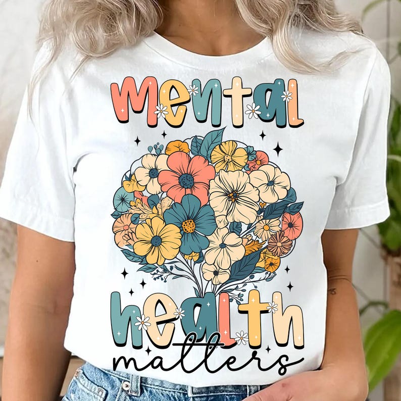 May include: White t-shirt with the words "mental health matters" in a colorful, retro font. A bouquet of flowers in shades of peach, blue, and yellow is centered on the shirt, with small star accents.