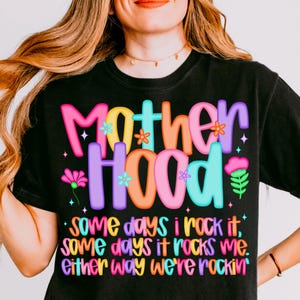 May include: Black t-shirt with the word "Motherhood" in colorful bubble letters. The shirt has the text "some days I rock it, some days it rocks me, either way we're rockin'" in a playful font. The shirt is a casual wear item.
