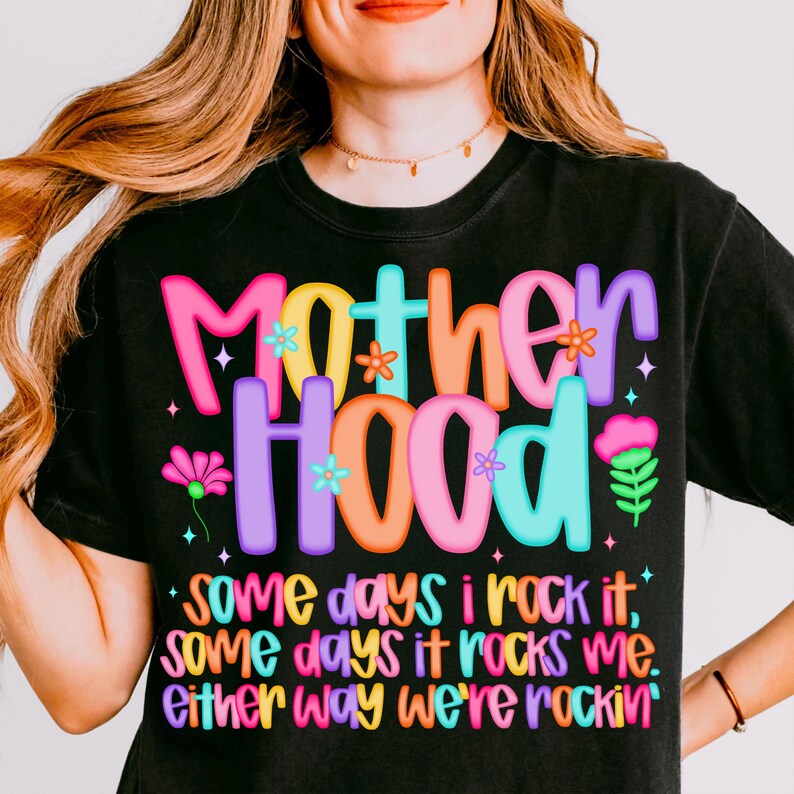 May include: Black t-shirt with the word "Motherhood" in colorful bubble letters. The shirt has the text "some days I rock it, some days it rocks me, either way we're rockin'" in a playful font. The shirt is a casual wear item.