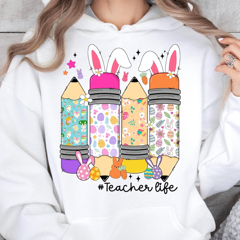 Op de afbeelding: Witte hoodie met een afbeelding van drie kleurrijke potloden versierd met paaseieren en konijnenoren. De tekst '#Teacher life' is onder de potloden gedrukt.