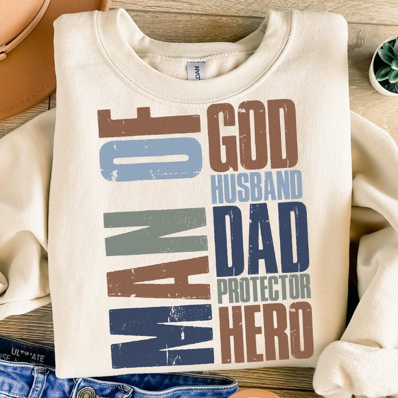 以下が含まれることがあります： クリーム色のスウェットシャツ。テキスト「MAN OF GOD, HUSBAND, DAD, PROTECTOR, HERO」が、使い古された茶色、青、グレーの文字でプリントされています。スウェットシャツはクルーネックと長袖です。テキストは左側と右側に縦に配置されています。