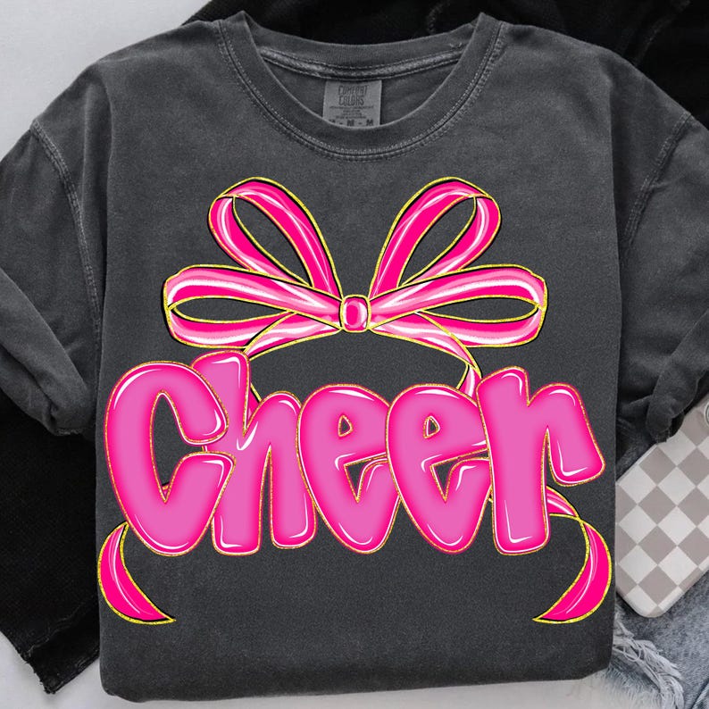 Puede incluir: Camiseta gris oscuro con un gran lazo rosa y la palabra "Cheer" en letras de burbujas. El lazo tiene un contorno dorado y la palabra "Cheer" es de color rosa brillante. La camiseta es de estilo cuello redondo.