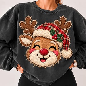 Può includere: Una felpa girocollo grigio antracite con una grande grafica testurizzata di una renna sorridente. La renna indossa un cappello da Babbo Natale a quadri rossi e verdi con un pon pon bianco e dettagli di agrifoglio. Il design ha bordi sfrangiati.
