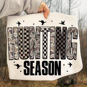 Puede incluir: Bolso tote color crema con las palabras "HUNTING SEASON" en un patr&oacute;n de camuflaje y cuadros. Siluetas de p&aacute;jaros y estrellas est&aacute;n dispersas alrededor del texto. El bolso es sostenido por una persona.