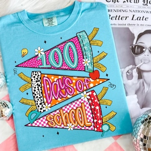 以下が含まれることがあります： 鮮やかな色のグラフィックデザインが施された水色のTシャツで、「100 Days of School」を祝っています。デザインは、「100」、「Days of」、「School」というテキストが鮮やかな色で描かれた3つのペナント型の要素と、花やリンゴなどの装飾要素が特徴です。