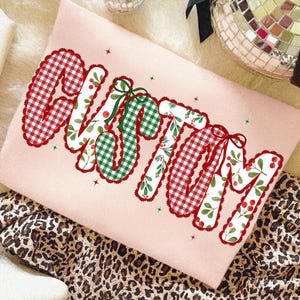 Pu&ograve; includere: Una felpa rosa chiaro con la scritta "CHRISTMAS" in un design festivo. Le lettere presentano motivi a quadri rossi e verdi, accenti floreali e fiocchi rossi. La felpa &egrave; esposta su un tessuto con stampa leopardata.