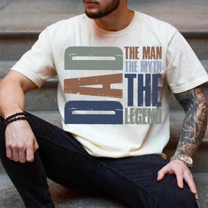 Peut inclure: T-shirt cr&egrave;me avec le texte "DAD" dans une police vieillie, au-dessus des mots "THE MAN THE MYTH THE LEGEND" dans un style assorti. Le t-shirt est port&eacute; par une personne assise sur des marches.