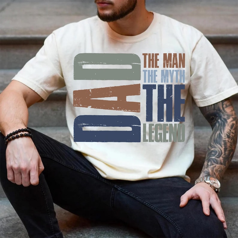 Peut inclure: T-shirt cr&egrave;me avec le texte "DAD" dans une police vieillie, au-dessus des mots "THE MAN THE MYTH THE LEGEND" dans un style assorti. Le t-shirt est port&eacute; par une personne assise sur des marches.
