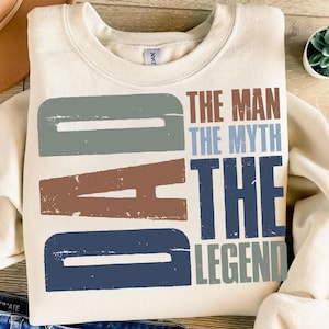 Peut inclure: Sweat-shirt cr&egrave;me avec le mot "DAD" en grandes lettres us&eacute;es bleues, marron et grises. Le texte "THE MAN THE MYTH THE LEGEND" est imprim&eacute; en marron, bleu et gris.