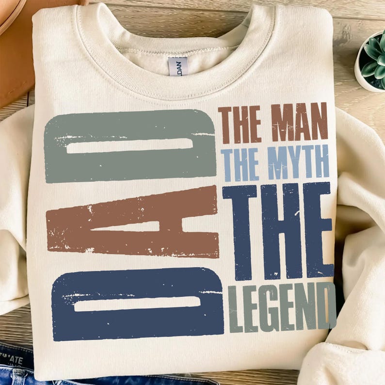 Peut inclure: Sweat-shirt cr&egrave;me avec le mot "DAD" en grandes lettres us&eacute;es bleues, marron et grises. Le texte "THE MAN THE MYTH THE LEGEND" est imprim&eacute; en marron, bleu et gris.