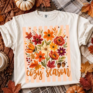 May include: A cream-colored short-sleeve t-shirt featuring a central graphic of autumn flowers, leaves, and two acorns over a light orange striped background. The text "cozy season" is printed in a cursive orange font below the floral design.