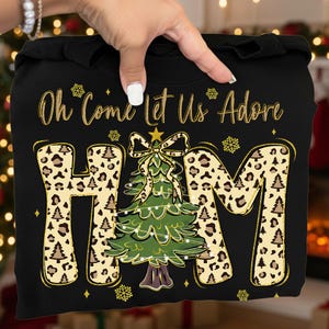 Può includere: Felpa nera con la scritta "Oh Come Let Us Adore Him". Le lettere "HIM" hanno un motivo leopardato con disegni di alberi di Natale. Un albero di Natale verde con un fiocco è al centro.