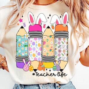 Op de afbeelding: Een wit T-shirt met een afbeelding van drie kleurrijke potloden met konijnenoren en paasei-patronen. De tekst "#Teacher life" is onder de potloden gedrukt.