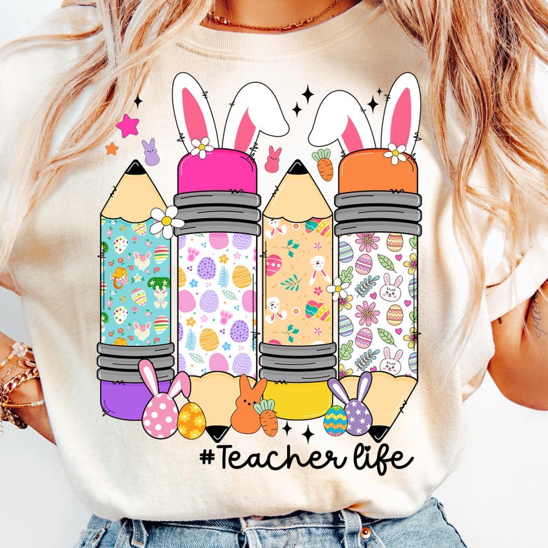 Op de afbeelding: Een wit T-shirt met een afbeelding van drie kleurrijke potloden met konijnenoren en paasei-patronen. De tekst "#Teacher life" is onder de potloden gedrukt.