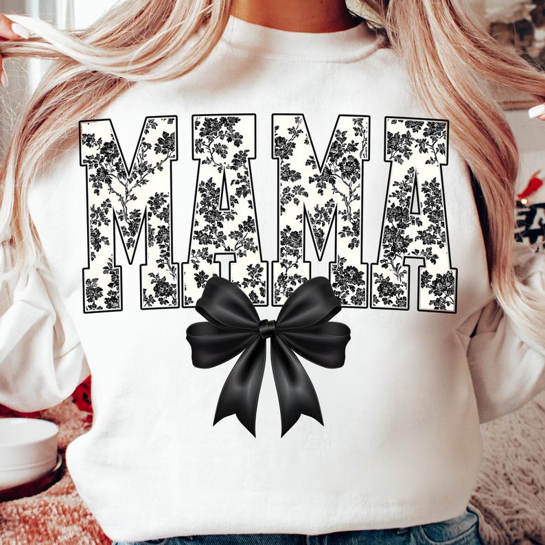May include: White sweatshirt with the word "MAMA" in large, black floral print letters. A black bow is centered below the word. The sweatshirt is a crew neck style.