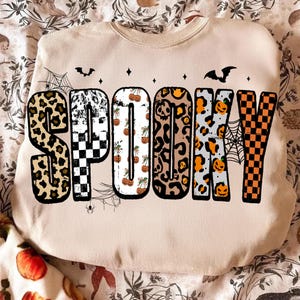 May include: Beige sweatshirt with the word "SPOOKY" in large, patterned letters. The letters feature leopard print, checkerboard, pumpkin, and spiderweb designs, with bat and star accents. A festive Halloween-themed garment.