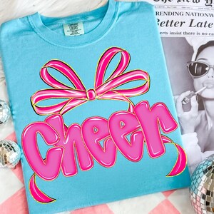 Puede incluir: Camiseta azul claro con un lazo rosa y la palabra "Cheer" en letras de burbujas. La camiseta est&aacute; hecha de un material suave, ideal para animadoras o uso diario.