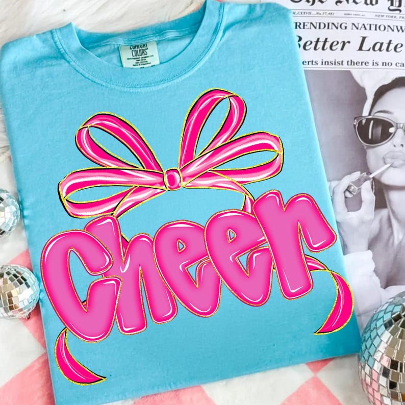 Puede incluir: Camiseta azul claro con un lazo rosa y la palabra "Cheer" en letras de burbujas. La camiseta est&aacute; hecha de un material suave, ideal para animadoras o uso diario.