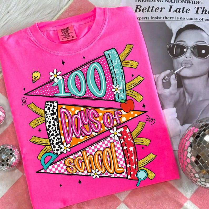 以下が含まれることがあります： 鮮やかなピンク色のTシャツ。カラフルなグラフィックで「100 Days of School」を祝っています。デザインには、テキスト、花、リンゴ、定規が描かれたペナントバナーが含まれています。柔らかい素材で作られたシャツです。