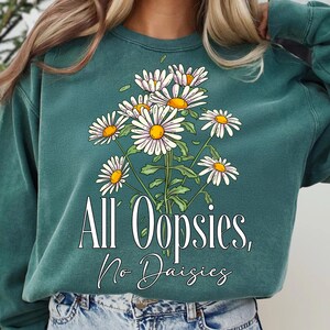 K&ouml;nnte beinhalten: T&uuml;rkisgr&uuml;nes Sweatshirt mit einem floralen G&auml;nsebl&uuml;mchen-Design. Die Grafik zeigt wei&szlig;e G&auml;nsebl&uuml;mchen mit gelben Zentren und gr&uuml;nen Stielen. Der Text "All Oopsies, No Daisies" ist darunter in Wei&szlig; gedruckt.