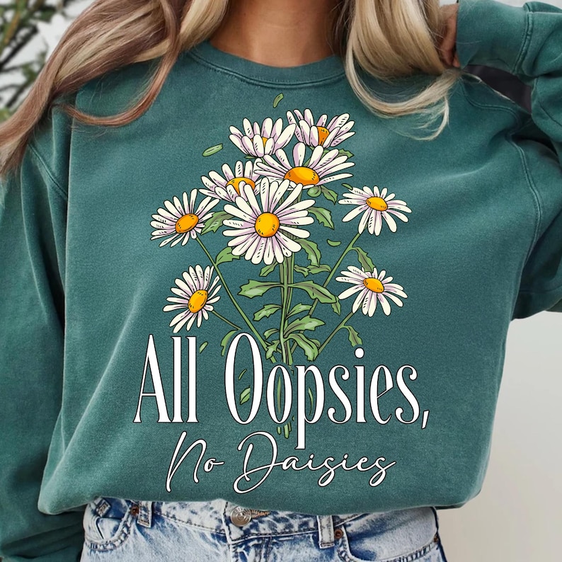 K&ouml;nnte beinhalten: T&uuml;rkisgr&uuml;nes Sweatshirt mit einem floralen G&auml;nsebl&uuml;mchen-Design. Die Grafik zeigt wei&szlig;e G&auml;nsebl&uuml;mchen mit gelben Zentren und gr&uuml;nen Stielen. Der Text "All Oopsies, No Daisies" ist darunter in Wei&szlig; gedruckt.