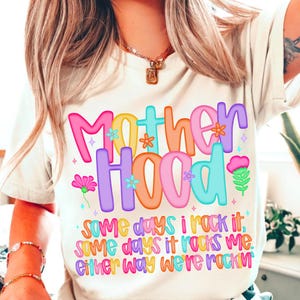 May include: A cream-colored t-shirt with the word "Motherhood" in colorful bubble letters. The shirt also features the text "Some days I rock it, some days it rocks me. Either way we're rockin".