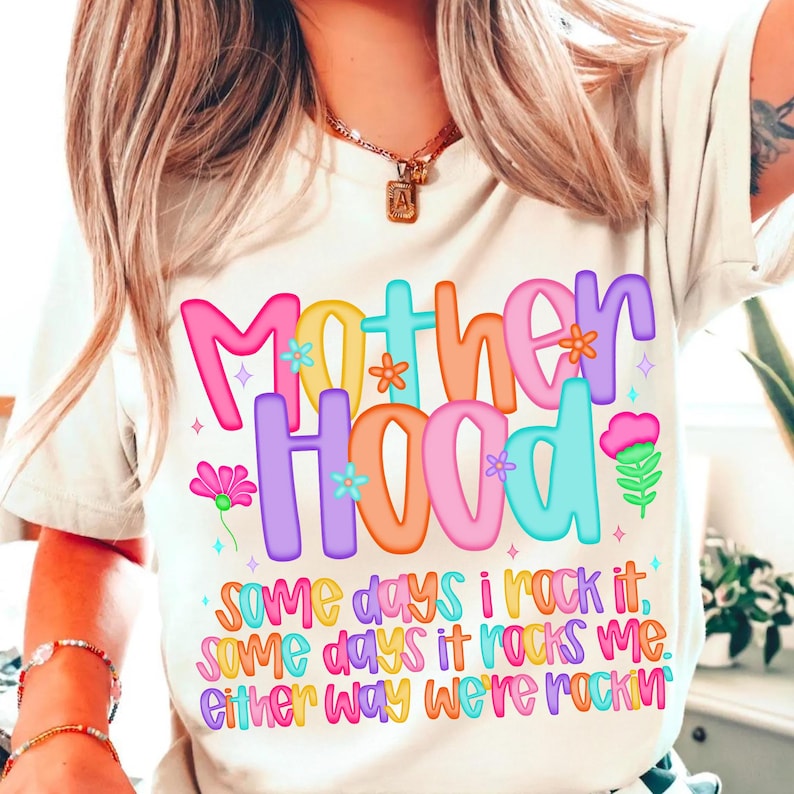 May include: A cream-colored t-shirt with the word "Motherhood" in colorful bubble letters. The shirt also features the text "Some days I rock it, some days it rocks me. Either way we're rockin".