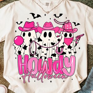 May include: Cream-colored t-shirt featuring a Halloween-themed graphic. The design includes three ghost figures wearing pink cowboy hats and bandanas, with a lasso and a toy gun. The text "Howdy Halloween" is in pink.
