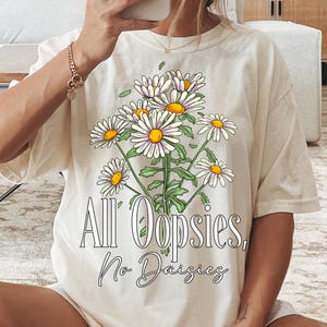 K&ouml;nnte beinhalten: Cremefarbenes T-Shirt mit einem G&auml;nsebl&uuml;mchen-Bouquet-Motiv mit wei&szlig;en Bl&uuml;tenbl&auml;ttern und gelben Zentren. Der Text "All Oopsies, No Daisies" ist darunter in einer verspielten Schriftart aufgedruckt. Das Shirt hat eine lockere Passform.